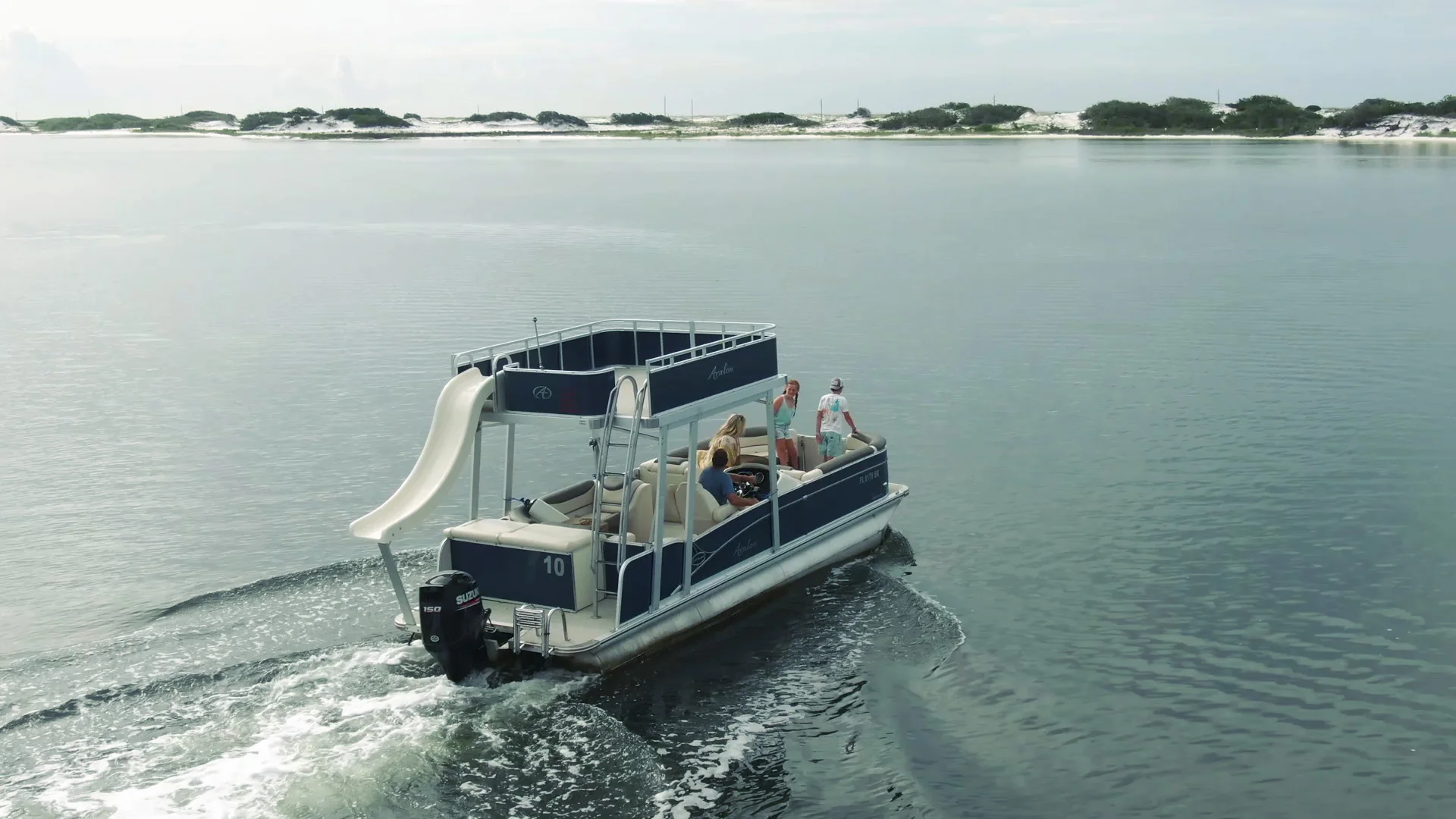 double-decker-pontoon-boat-rental-slide-navarre-florida A blue Avalon double-decker Pontoon Boat with a white Slide coming off the back idles across the glassy water of Navarre's intercoastal waterway