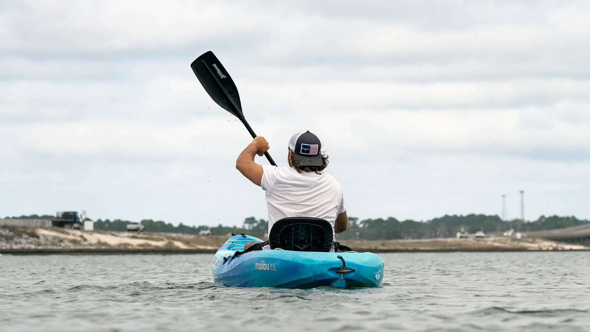 navarre-beach-kayak-rentals A man in a white shirt and backwards hat paddles a lone blue kayak across the Navarre intercoastal waterway