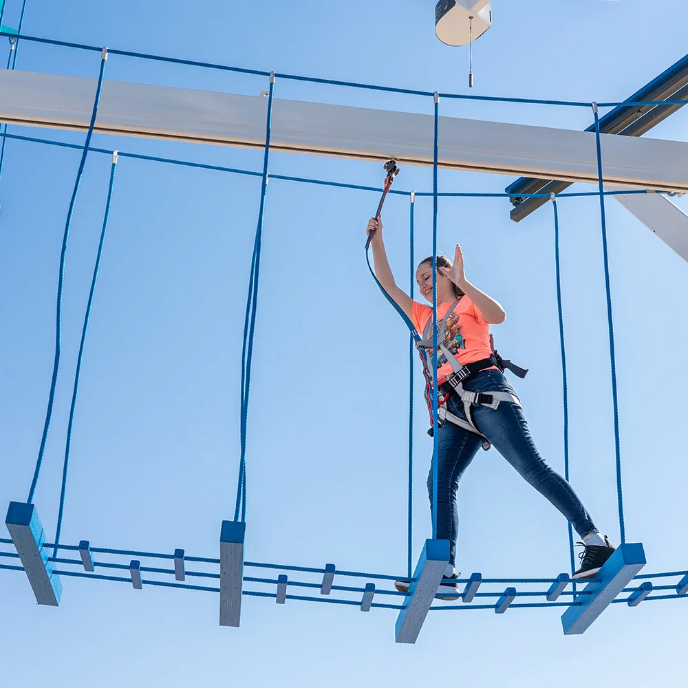 A girl in an orange shirt and blue jeans navigates her way across on of the many rope bridges on Navarre Family Watersports' Ropes Course